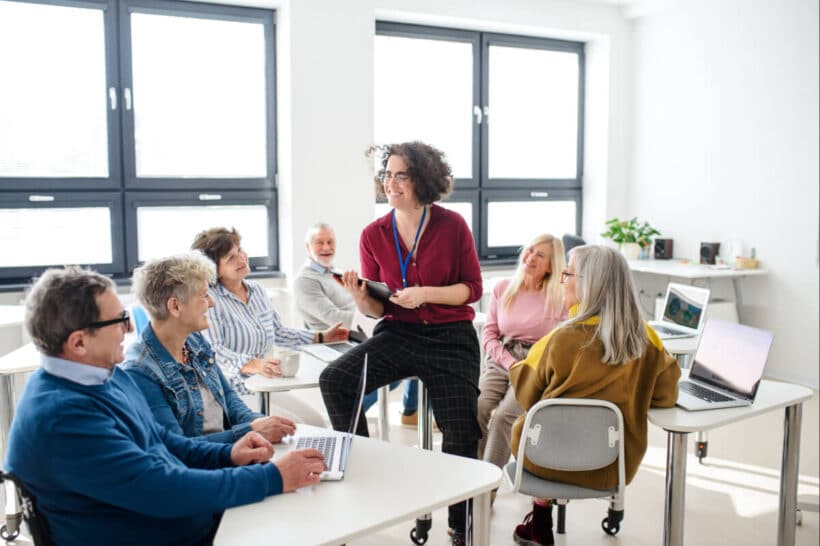 Gruppe Erwachsener in hellem Seminarraum; Kursleitung mit Tablet sitzt auf einem Tisch und moderiert, Teilnehmende mit Laptops hören und diskutieren.